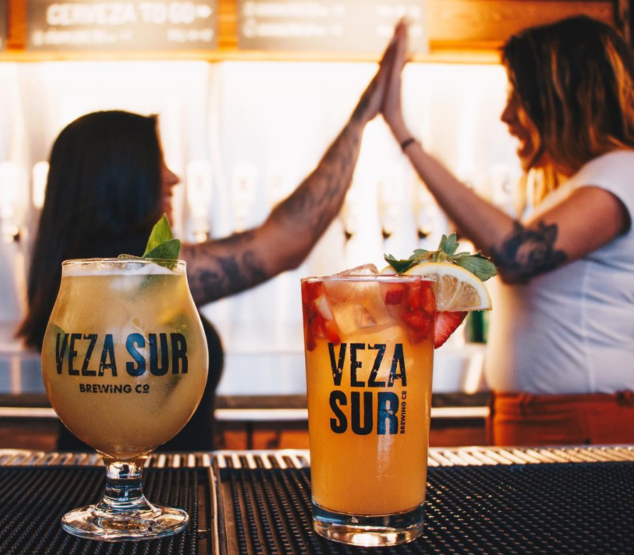 image of two drinks served at Veza Sur, a bar in Miami. In the background, waitresses highfive. 