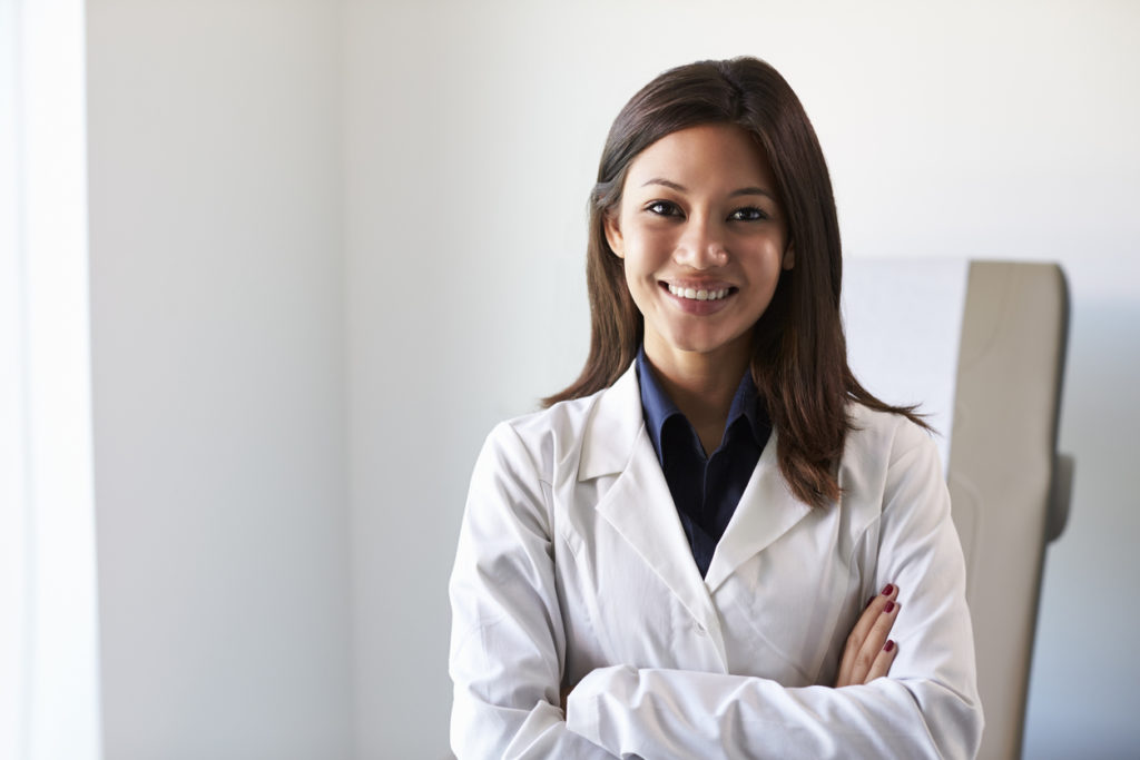 Portrait Of Female Doctor Wearing White Coat In Exam Room